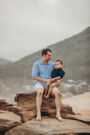 A father sits cuddling his son on rocks at the beach