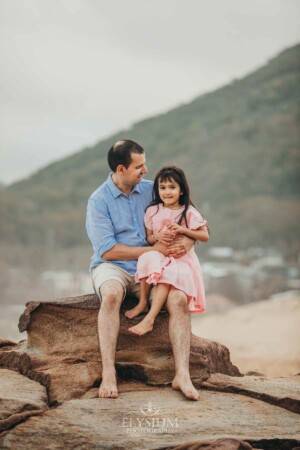 A father sits cuddling his daughter on rocks at the beach