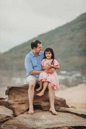 A father sits cuddling his daughter on rocks at the beach