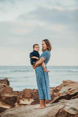 A mother stands cuddling her son on rocks at the beach