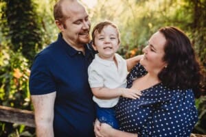 Parents cuddle their little boy between them as they stand amongst the trees