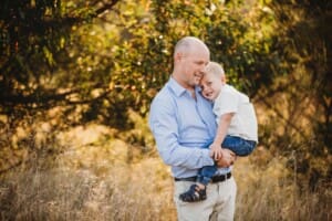A dad cuddles his son in a long grass field at sunset