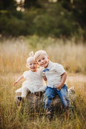 2 children sit together on a log in a grassy field