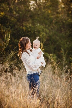 A mother holds her baby girl in a long grass field at sunset