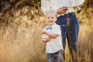 A little boy stands in long grass at sunset with his mum