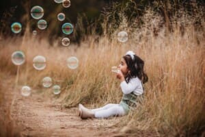 A little girl sits in long grass and blows bubbles