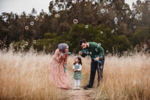 Parents stand in long grass and blow bubbles for their daughter