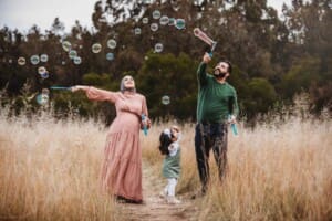 Parents stand in long grass and blow bubbles for their daughter