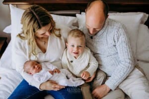 A little boy sits on a bed with his parents as they hold their newborn baby
