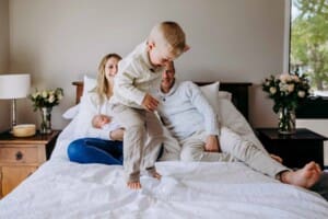 A little boy jumps on the bed with his parents sitting behind him