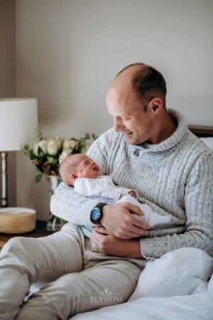 A dad sits on a bed and holds his newborn baby in his arms