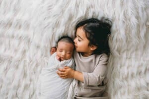 A newborn in a white wrap gets kisses from his big sister
