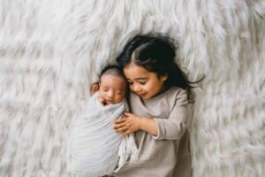 A newborn in a white wrap gets hugs from his big sister