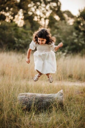 A little girl jumps off a log into long grass at sunset