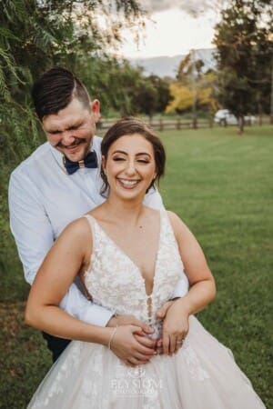 Gledswood Wedding - a groom cuddles his bride as they stand under a pepper tree at sunset