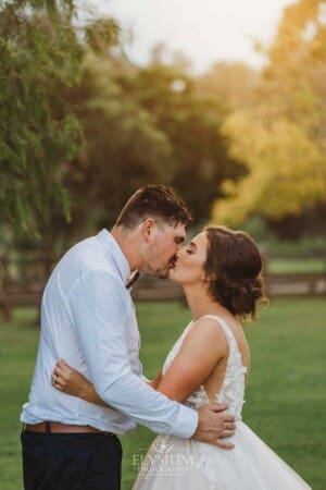 Gledswood Wedding - a groom kisses his bride as they stand under a pepper tree at sunset