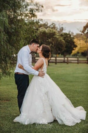 Gledswood Wedding - a groom kisses his bride as they stand under a pepper tree at sunset