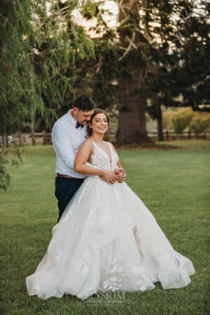 Gledswood Wedding - a groom cuddles his bride as they stand under a pepper tree at sunset