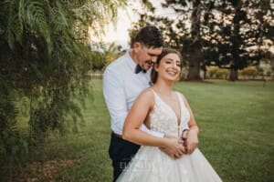 Gledswood Wedding - a groom cuddles his bride as they stand under a pepper tree at sunset