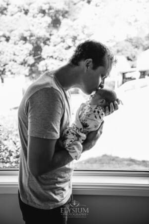 A father kisses his newborn baby on the head standing against a bright window