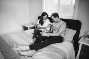 A couple sit on a bed holding their newborn between them