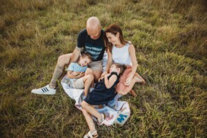 Parents tickle their children as they sit on a rug in a grassy field at sunset