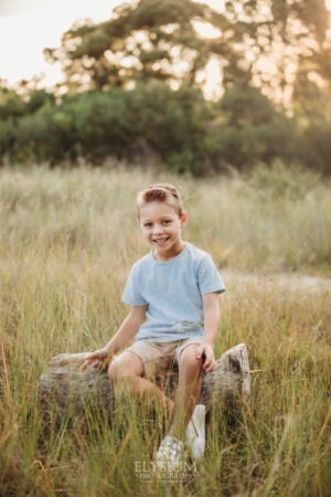 A little boy sitting on a log in a grassy field at sunset