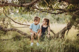 Two children sit on a low tree branch in a grassy field at sunset