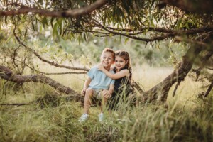 Two children sit on a low tree branch in a grassy field at sunset