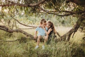 Two children sit on a low tree branch in a grassy field at sunset