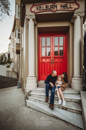 Baby photography - A family cuddle their baby girl as they sit in front of a red door