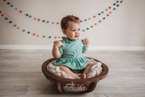 A baby girl wearing a green romper sits in a rustic wood bowl in a bright studio
