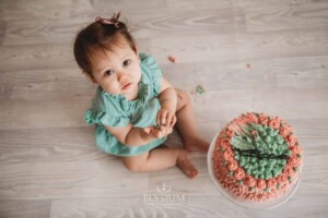 A baby girl sitting next to her birthday cake on a white studio floor