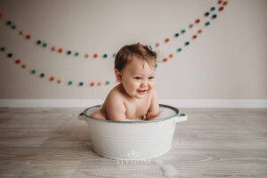 A baby girl sits in a white bath tub cleaning up after her cake smash session