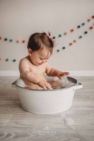 A baby girl splashing in a bubble bath after her cake smash session