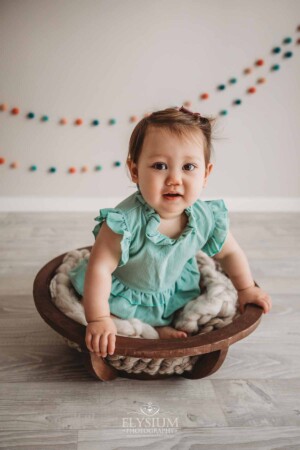 A baby girl wearing a green romper sits smiling in a wooden bowl in a bright studio