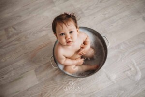A baby girl sitting in a vintage tub enjoys a bubble bath after her messy cake smash