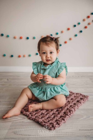 A baby girl wearing a green romper sits on a pink blanket with coloured bunting behind her