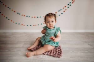 Baby girl wearing a green romper sitting on a pink rug with coloured bunting behind her