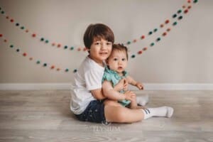 A kid holding his baby sister in his lap with coloured bunting behind them