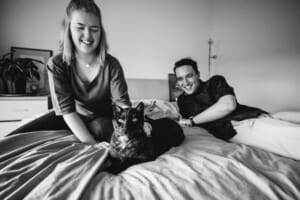 Engagement Photographer: a couple sit on a white bed with their black cat
