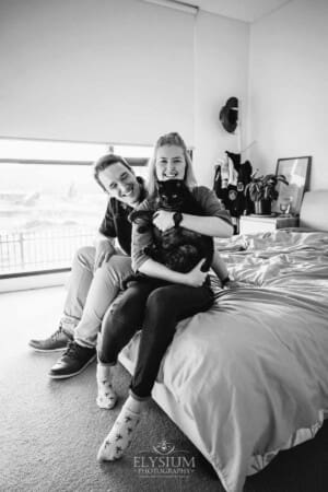 Engagement Photographer: a couple sit on a white bed with their black cat
