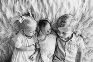 Siblings hold their newborn sister between them laying on a bed