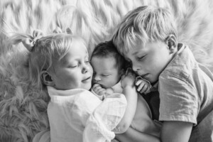 Siblings hold their newborn sister between them laying on a bed
