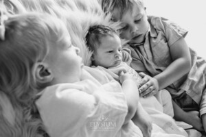 Siblings hold their newborn sister between them laying on a bed