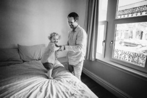 Newborn Photography: a little boy holds his dad's hands as he jumps on a bed