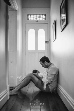 Newborn Photography: a father sits in a hallway with his baby in his lap