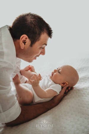 Newborn Photographer: a father hugs his baby boy laying on a white bed