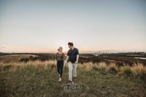 Engagement Photographer: a couple walk holding hands across a grassy hill at sunset
