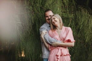 Engagement Photography: a couple stand cuddling against a green hedge